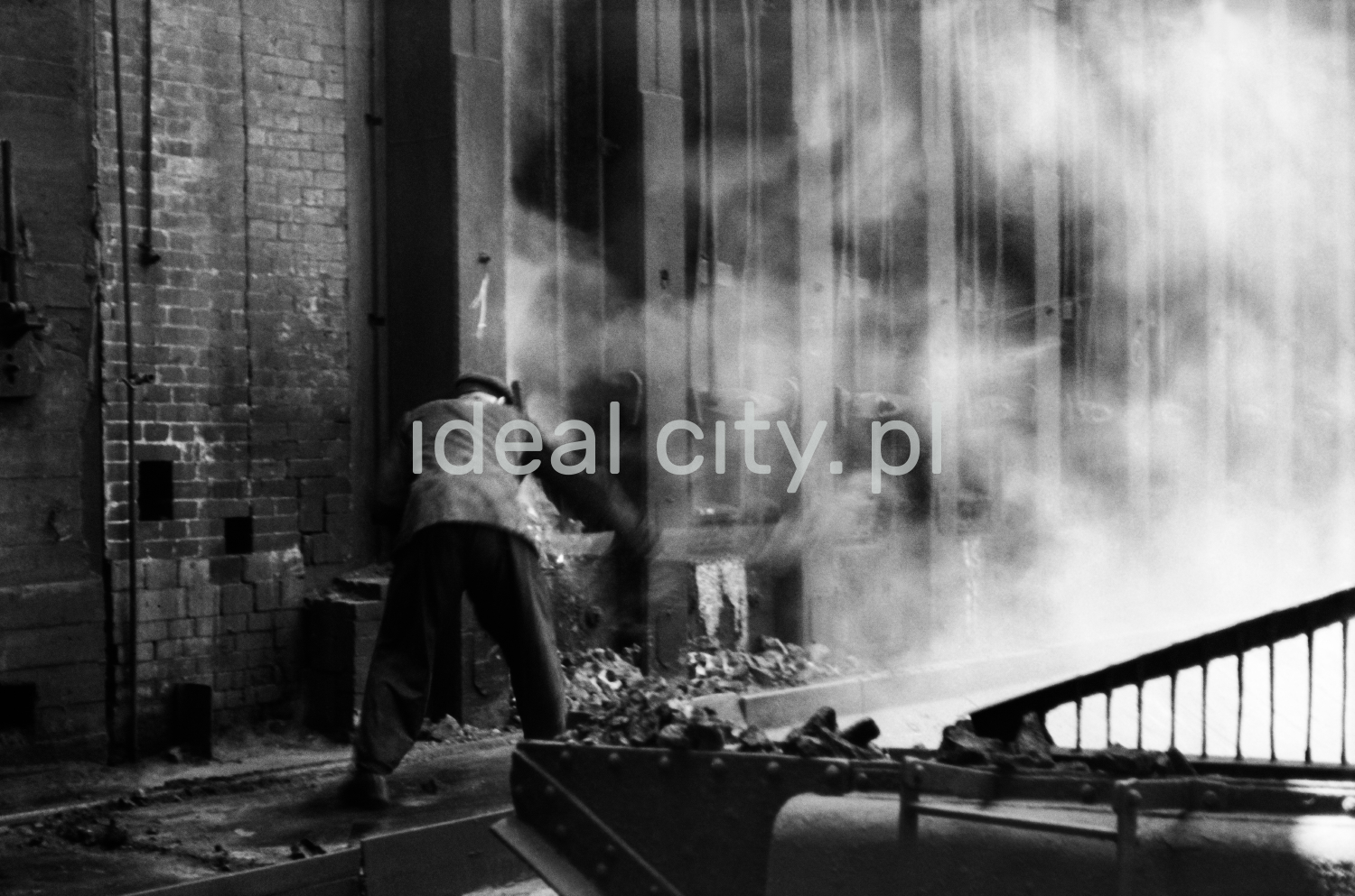 A man in work clothes uses a shovel to throw coke into a hole in a huge wall of a metal structure.