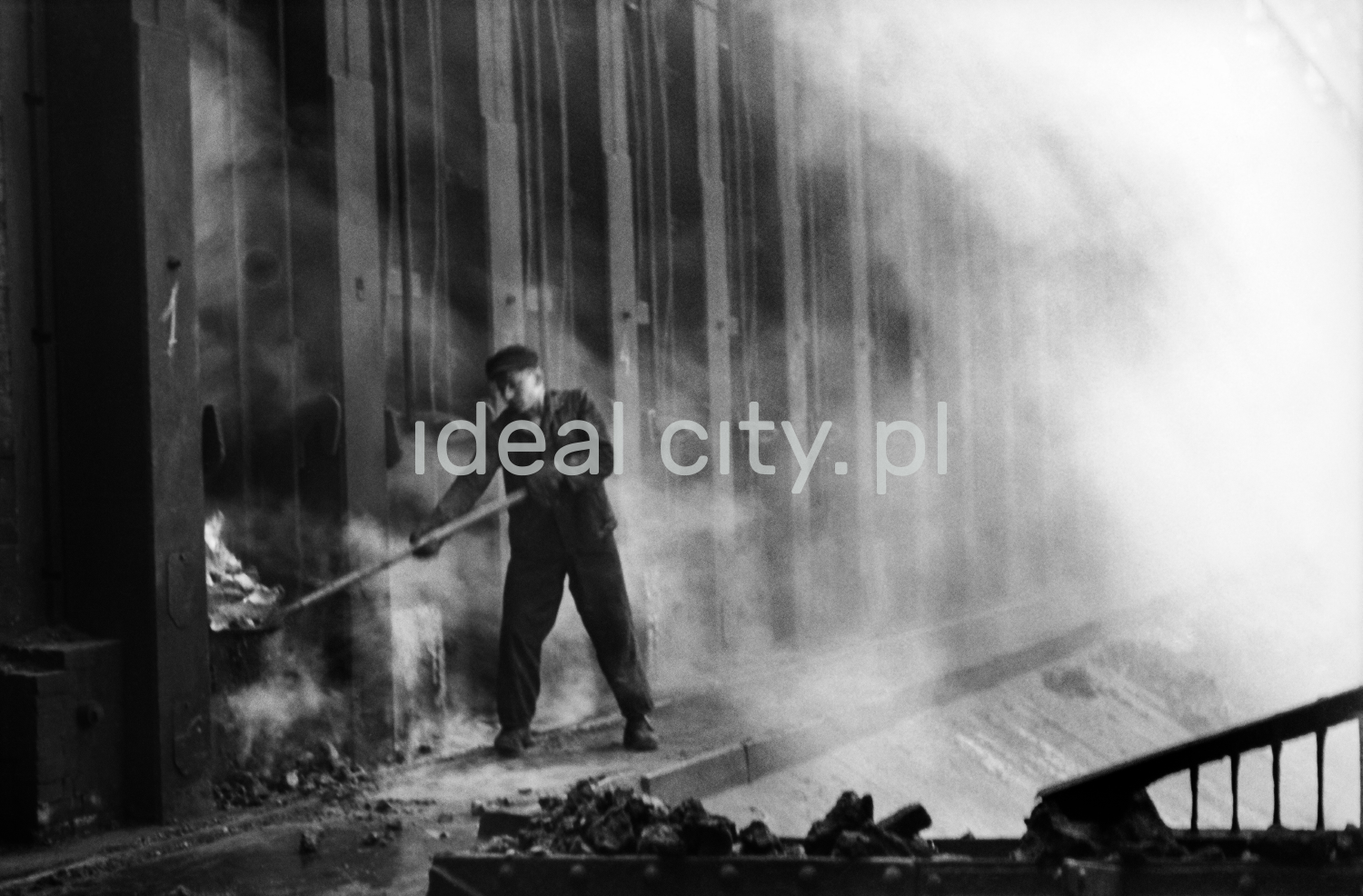 A man in work clothes uses a shovel to throw coke into a hole in a huge wall of a metal structure.