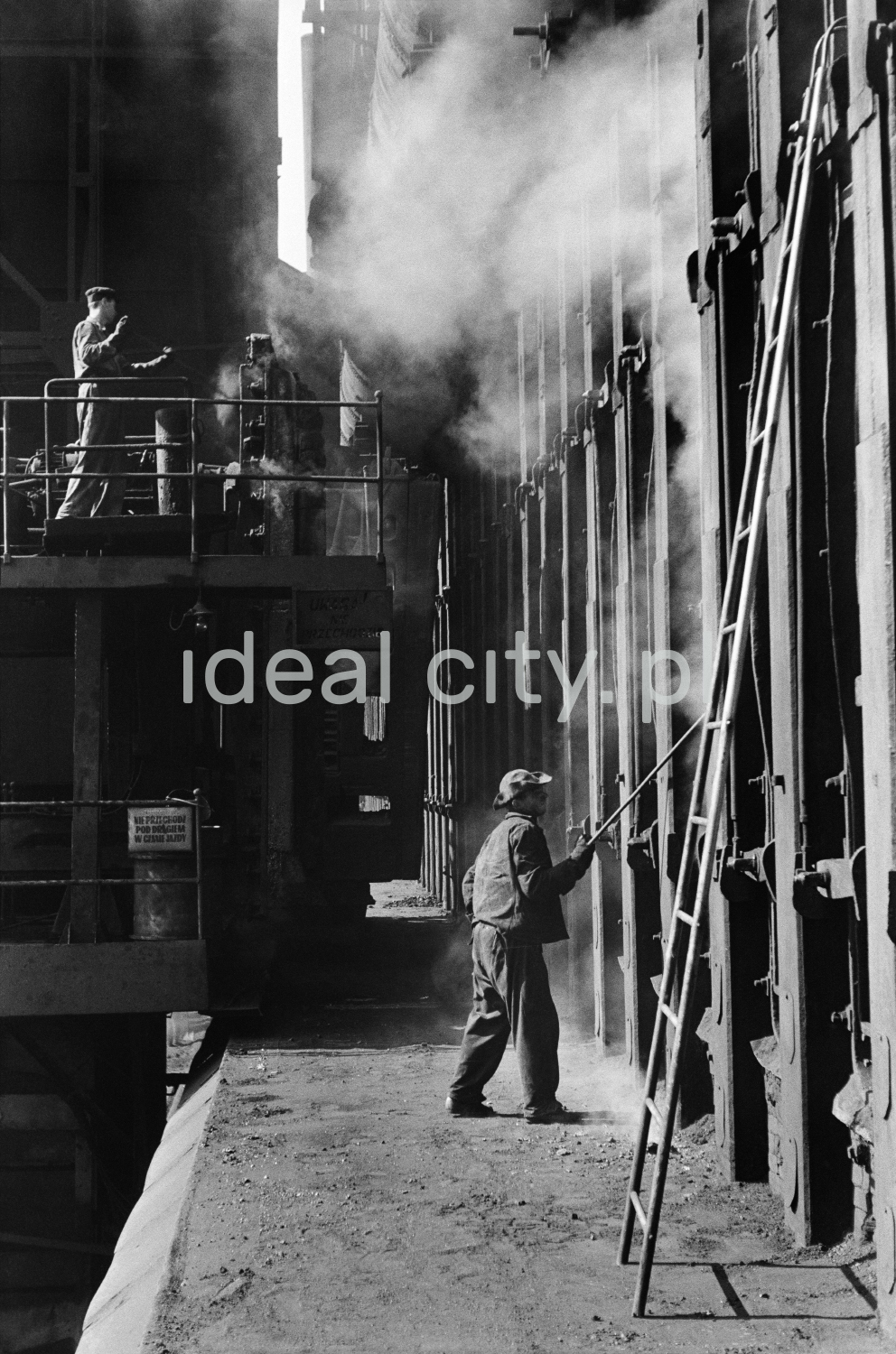 The steelworker inserts a long rod into a massive steel structure.