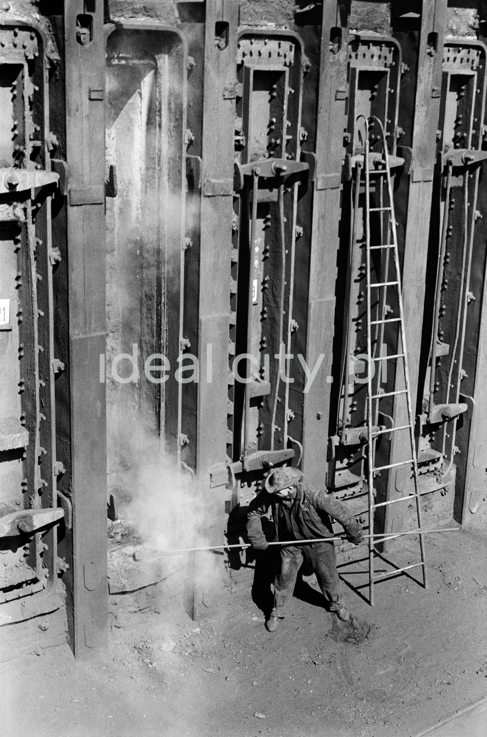 The steelworker inserts a long rod into a massive steel structure.