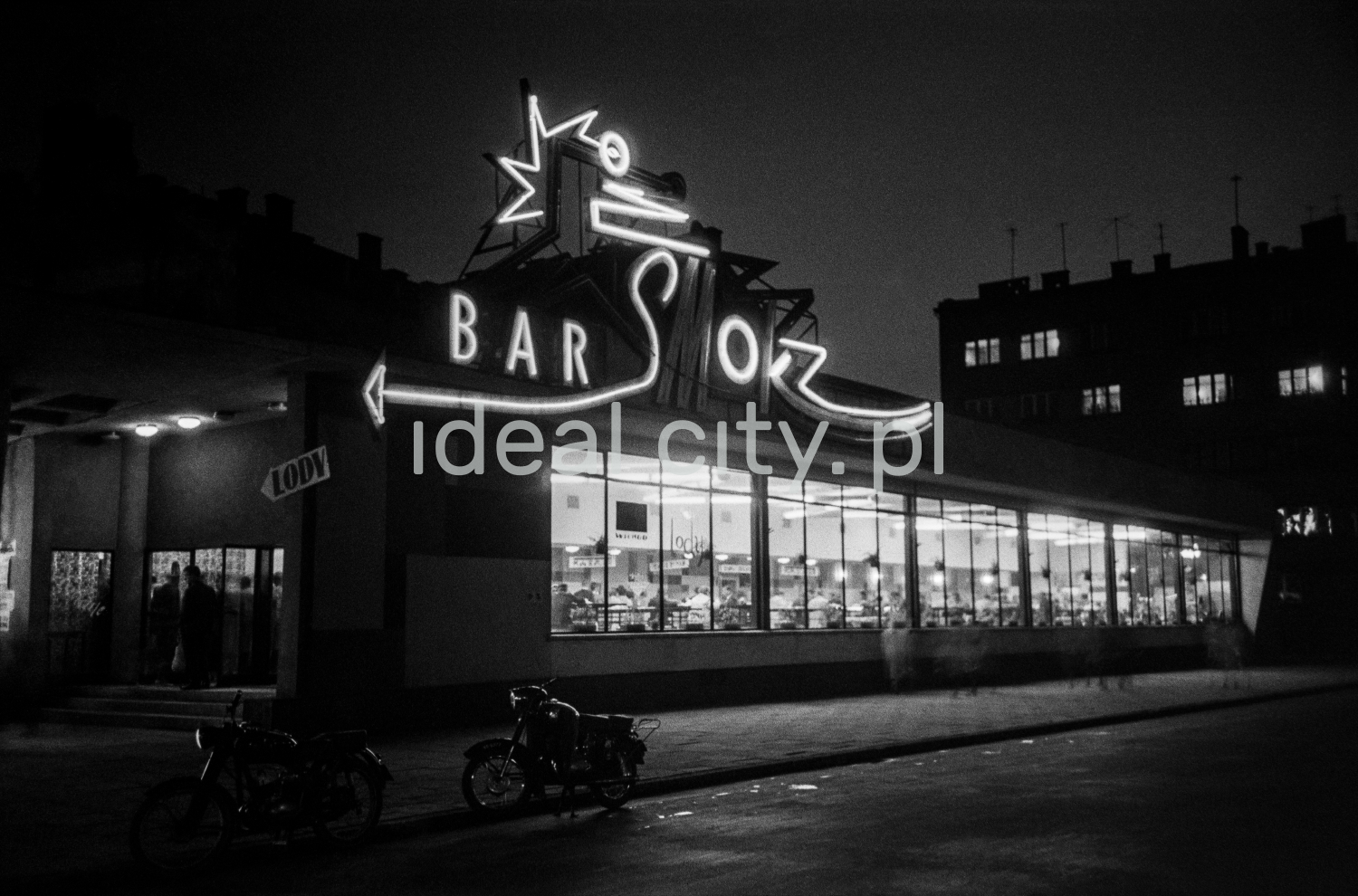 A night shot of a modernist lighted bar pavilion with a neon sign.