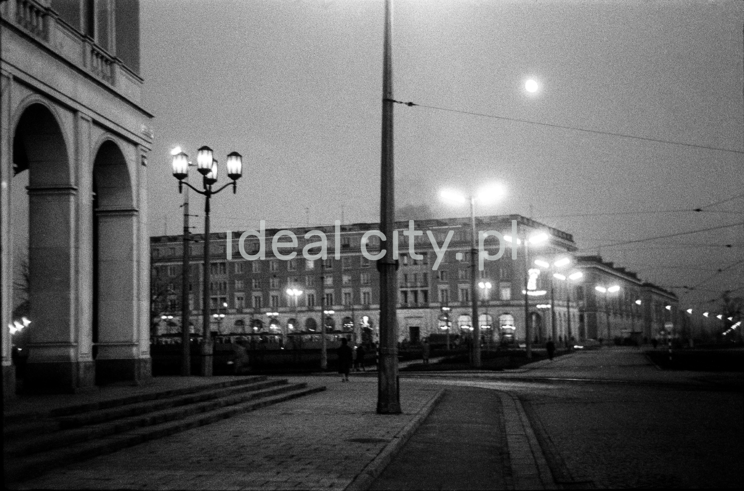 A night shot of a spacious, illuminated square with circular tram traffic, monumental residential buildings all around.