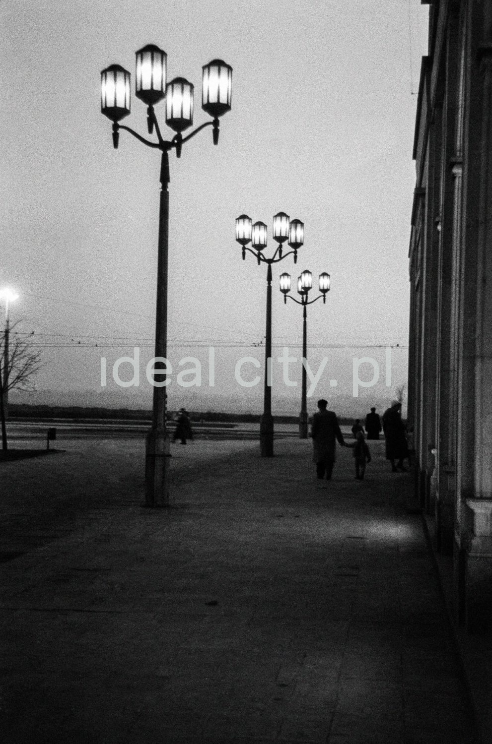 A night shot of a series of monumental lanterns illuminating the wide pavement along the building, visible figures of passers-by.