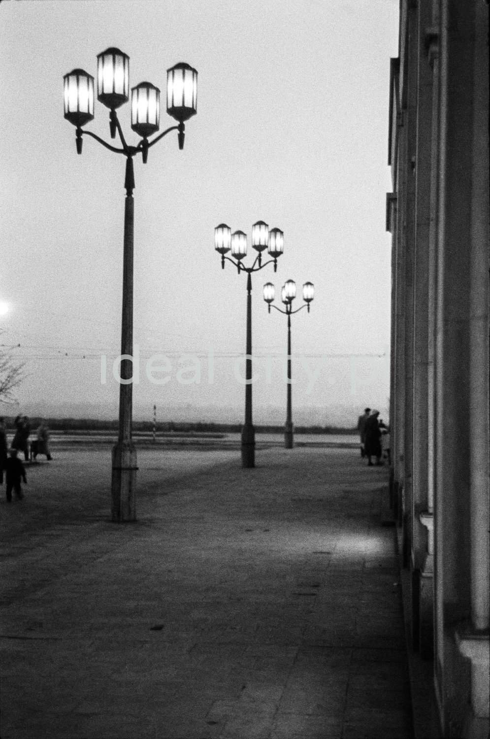 A night shot of a series of monumental lanterns illuminating the wide pavement along the building, visible figures of passers-by.
