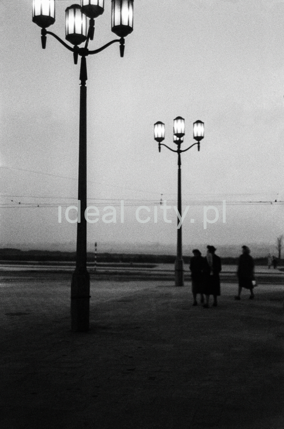A night shot of a series of monumental lanterns illuminating the wide pavement along the building, visible figures of passers-by.
