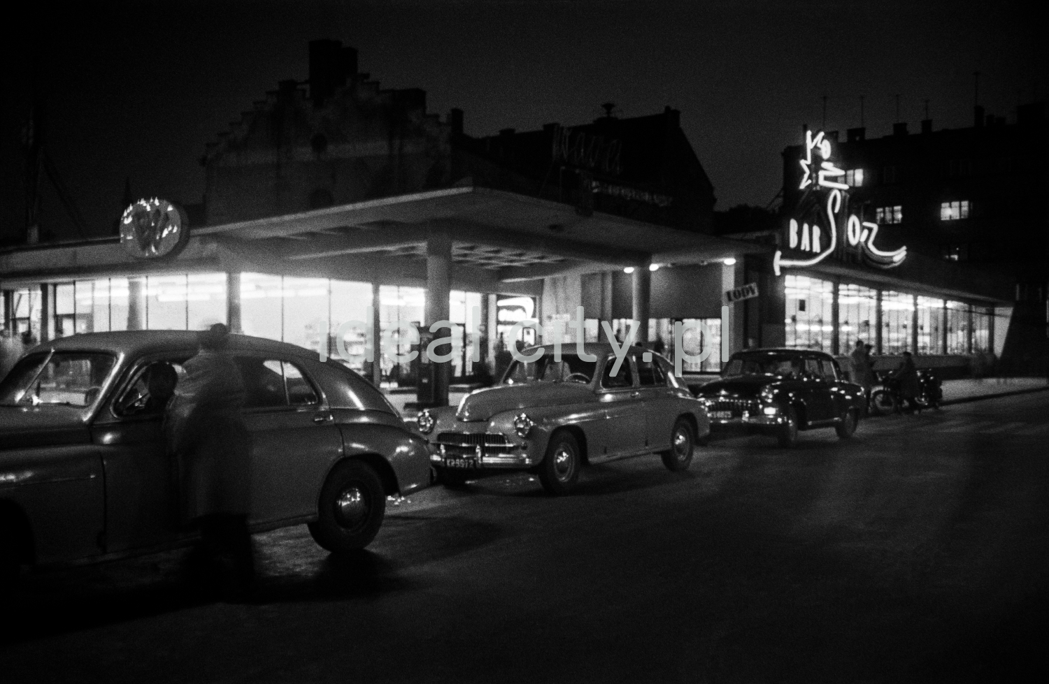A night shot of a modernist lighted bar pavilion with a neon sign.