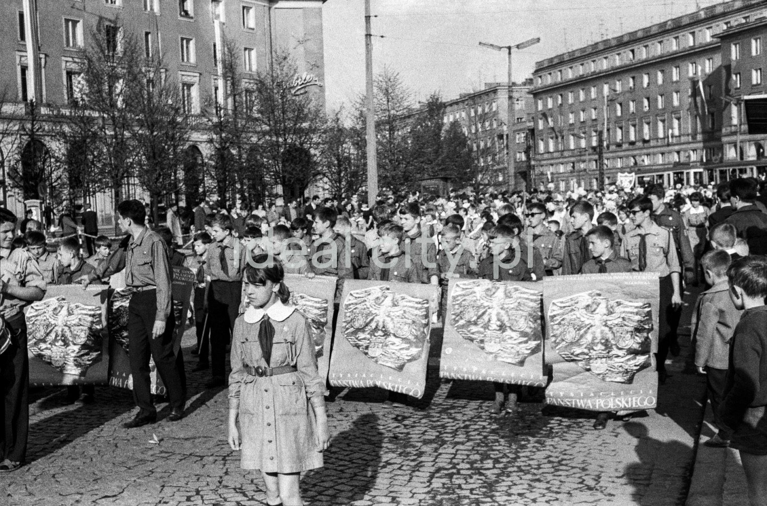 A view of the wide street where children are marching, with modernist apartment blocks in the background.