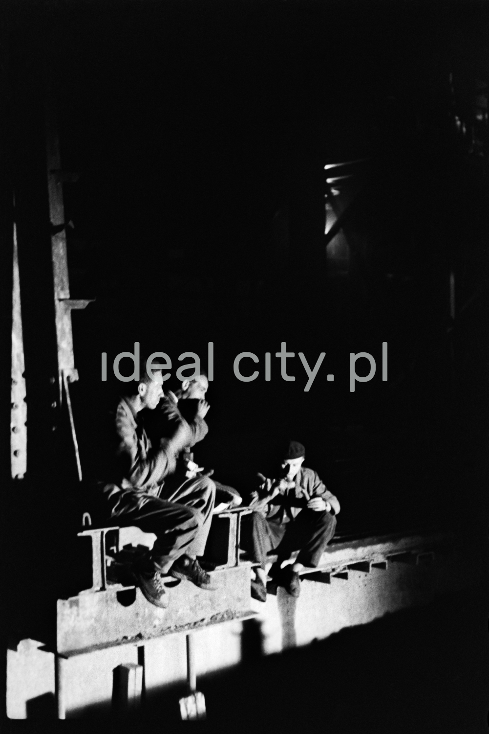 Men in working clothes on a steel beam placed at a height in a dark hall during a break.