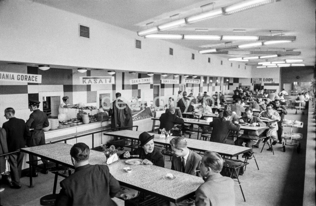 People in overcoats eat their meals by a row of openwork bar tables inside a bright, modernist pavilion.