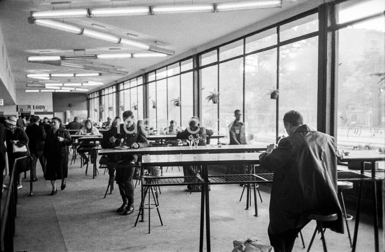 People in overcoats eat their meals by a row of openwork bar tables inside a bright, modernist pavilion.