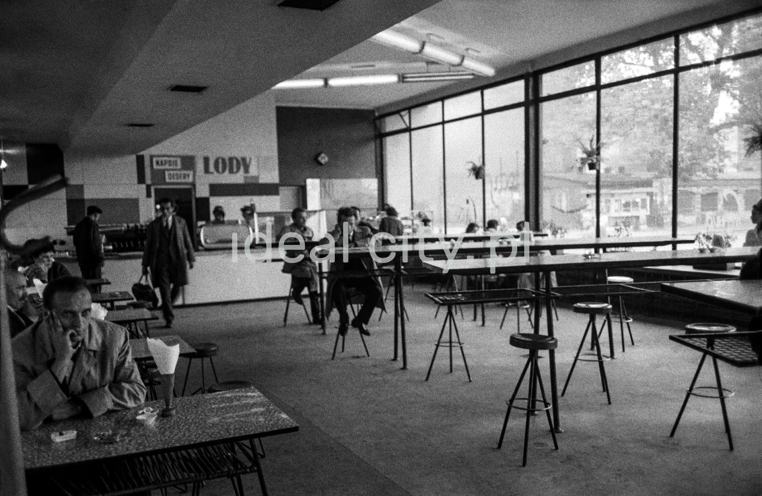 People in overcoats eat their meals by a row of openwork bar tables inside a bright, modernist pavilion.