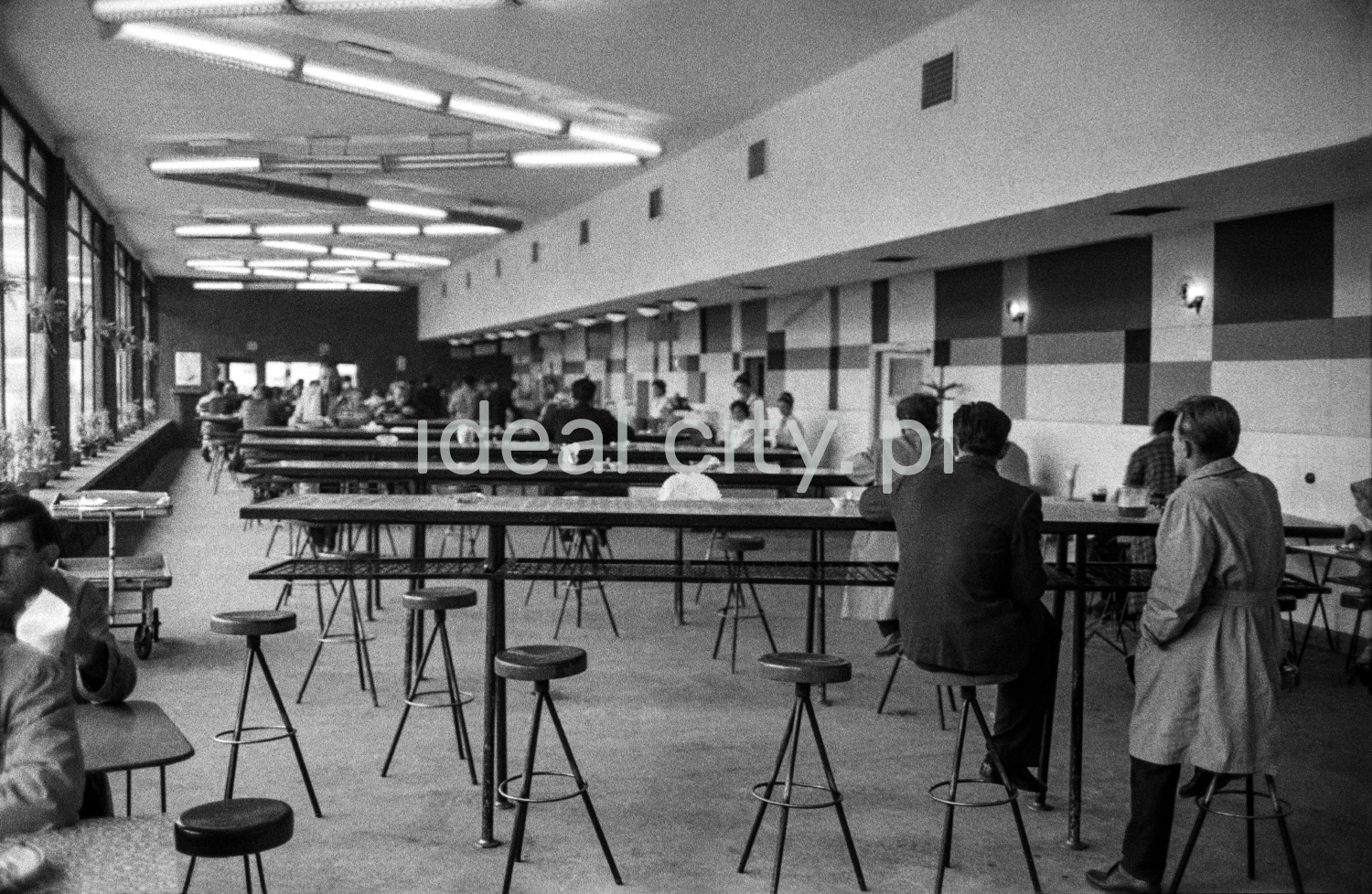 People in overcoats eat their meals by a row of openwork bar tables inside a bright, modernist pavilion.