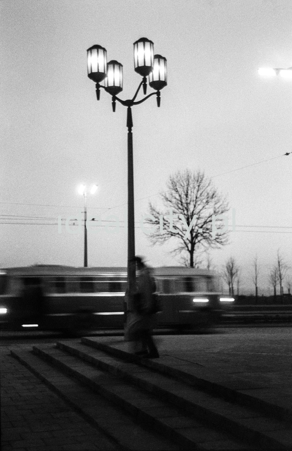A view of the monumental street lamp, visible silhouettes of passers-by and a bus passing in the background.