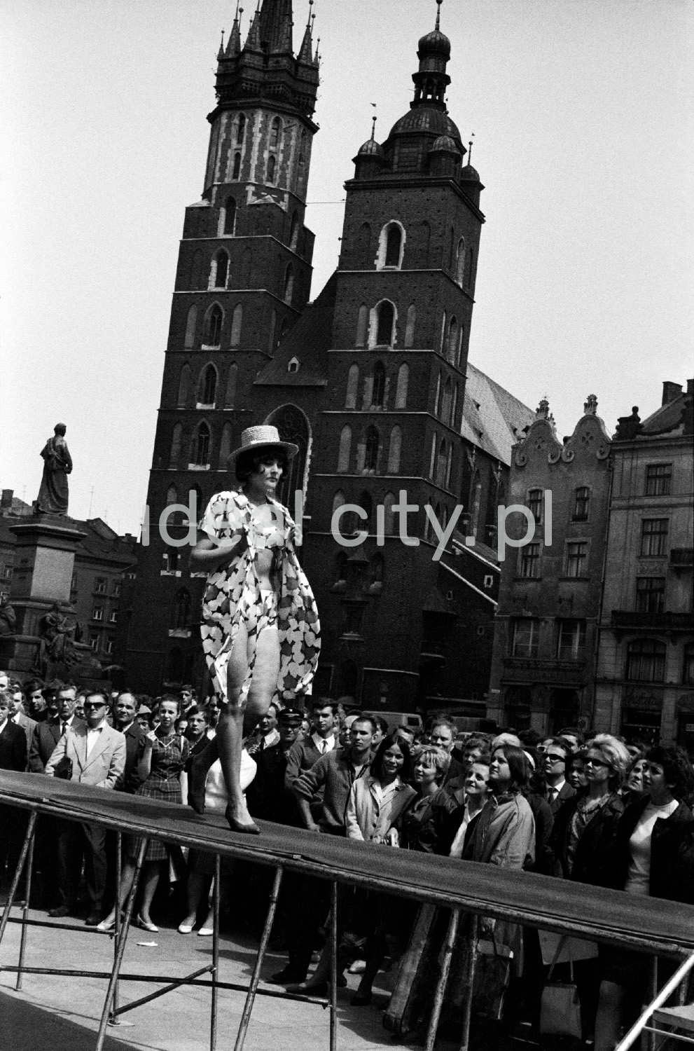 A woman in a dress and a hat is walking along the catwalk located in the town square surrounded by historic buildings, in the background a Gothic church with two towers.