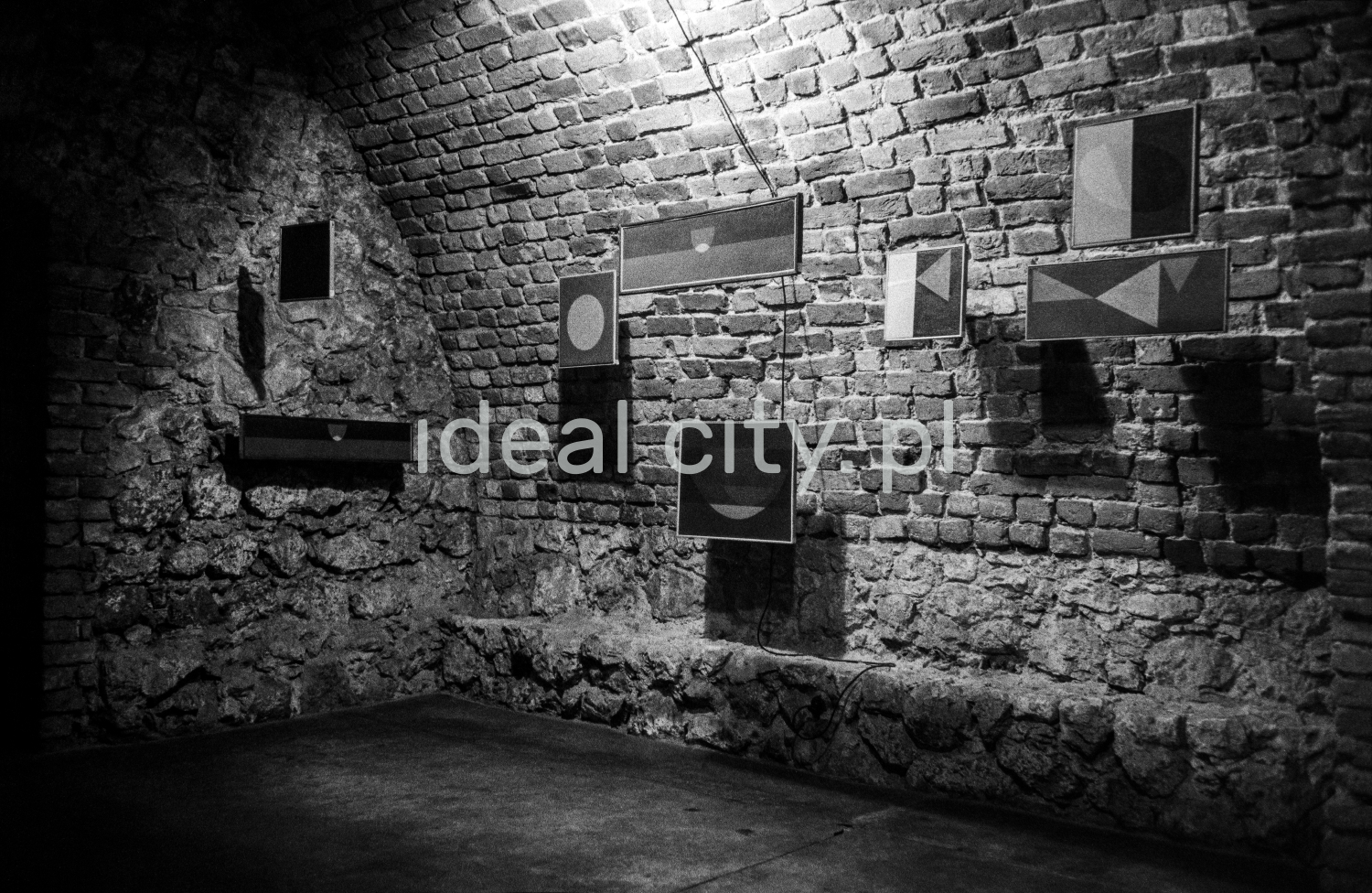 A view of a brick cellar with a semicircular vault, paintings on the walls illuminated by halogens.