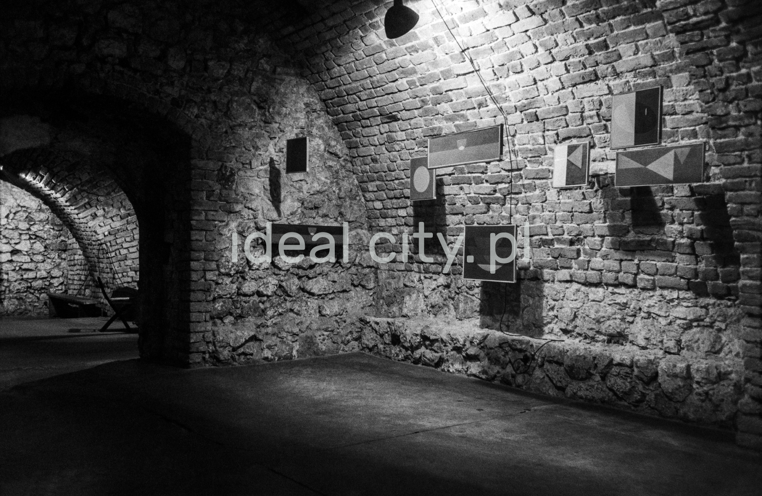 A view of a brick cellar with a semicircular vault, paintings on the walls illuminated by halogens.