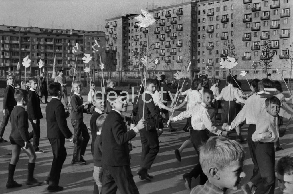 Primary school students in festive costumes walk in the procession holding pigeons cut out of cardboard in their hands. A sprawling apartment block in the background.