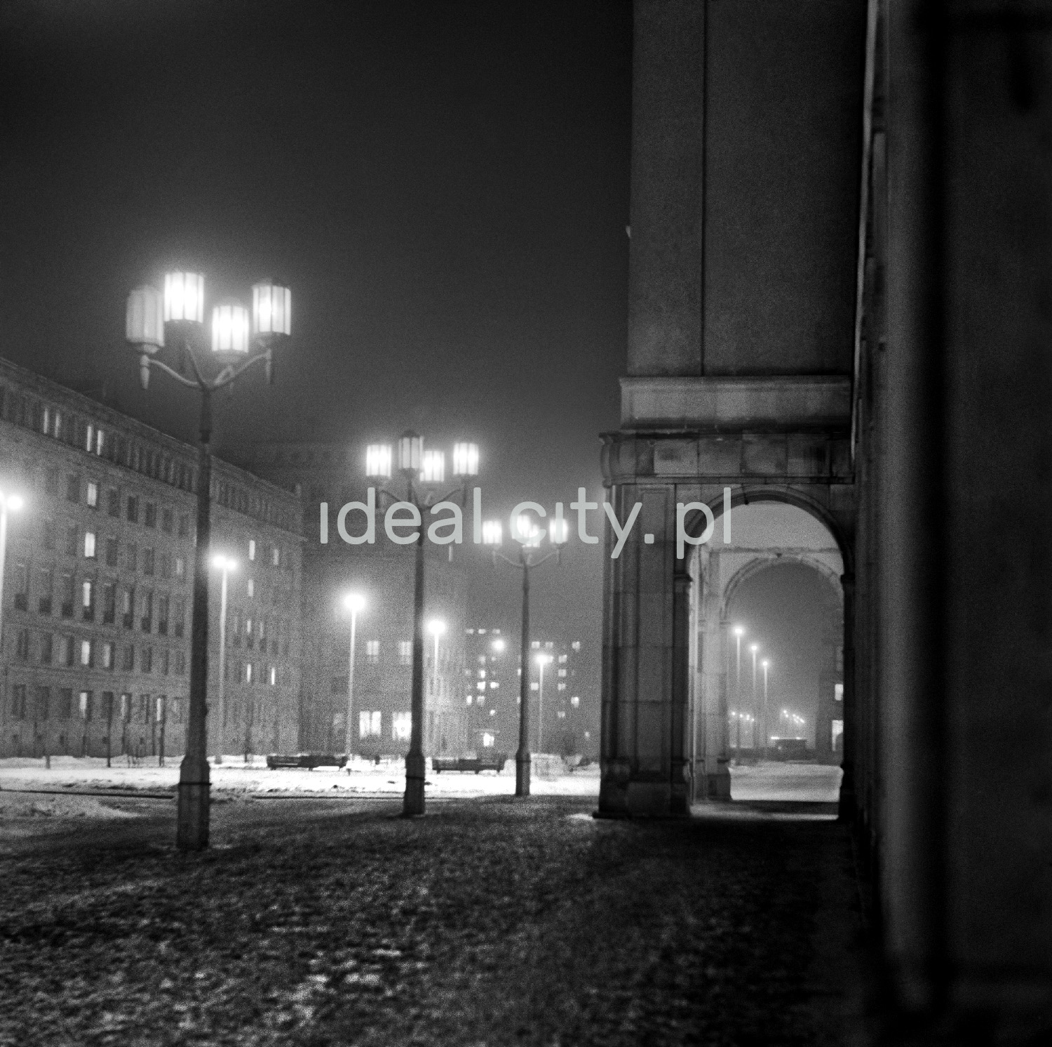 A view of a wide avenue lit by street lamps, with no car traffic, on the right there is an arcade under a monumental residential building.