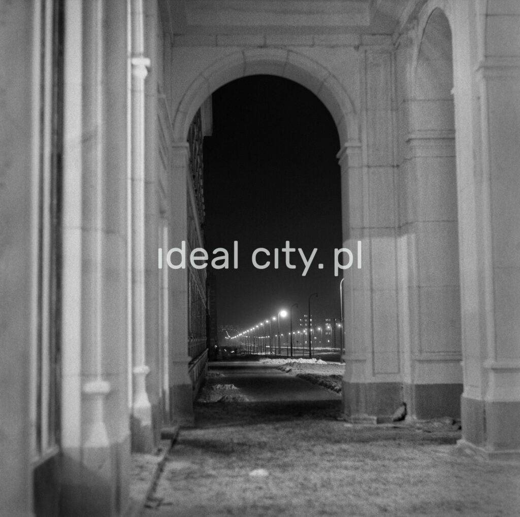 A night-time perspective of a street lit by street lamps seen from under monumental arcades.