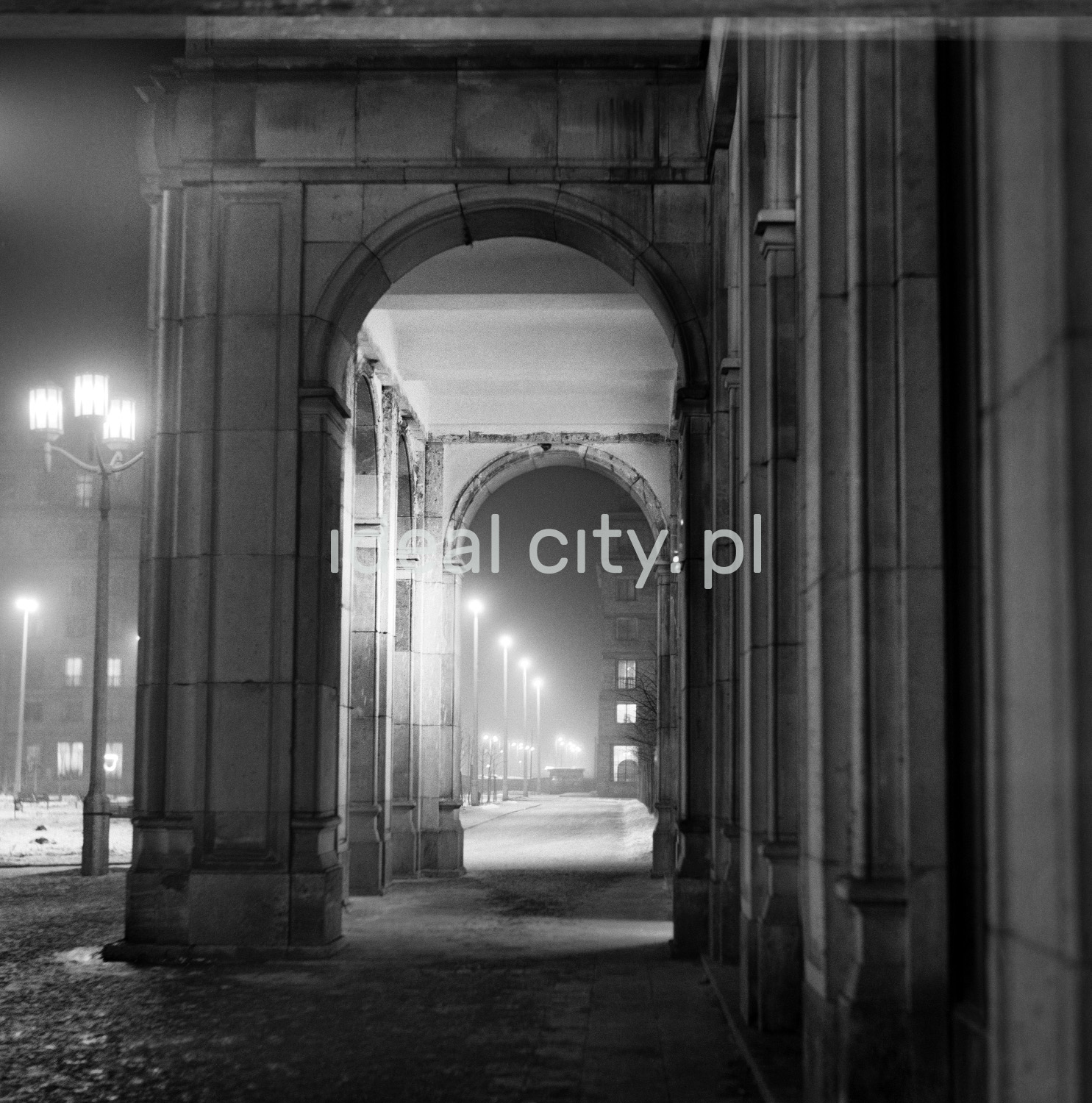 A view of a wide avenue lit by street lamps, with no car traffic, on the right there is an arcade under a monumental residential building.