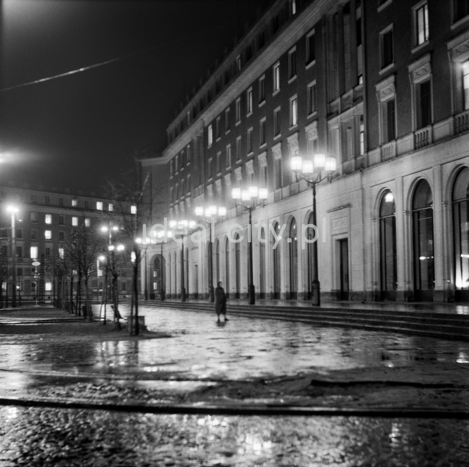 A view of a wide square lit by street lamps, with no car traffic, on the right there is an arcade under a monumental residential building.