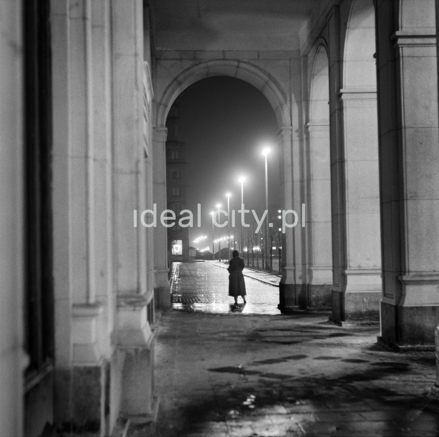 A night-time perspective of a street lit by street lamps seen from under monumental arcades. In the center, there is a silhouette of a woman in a coat.