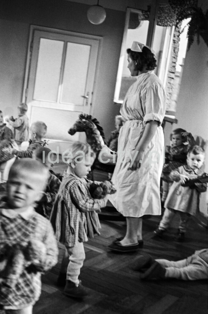A babysitter in a bright apron stands among the playing children.