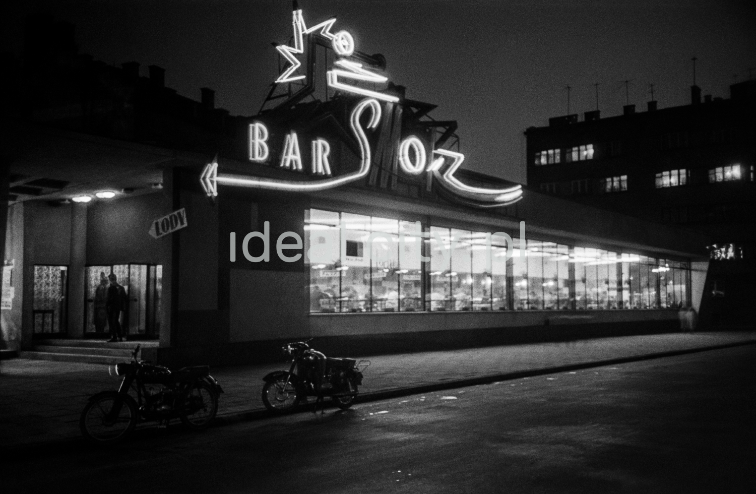 A night shot of a modernist lighted bar pavilion with a neon sign.