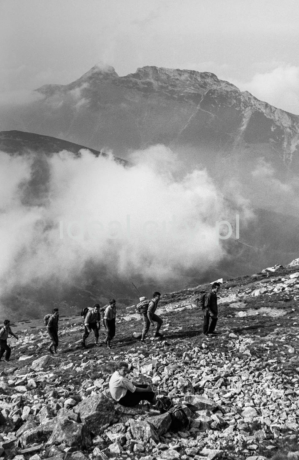 Tourists with backpacks are walking along the rocky trail, with the perspective of the Tatra peaks in the background.