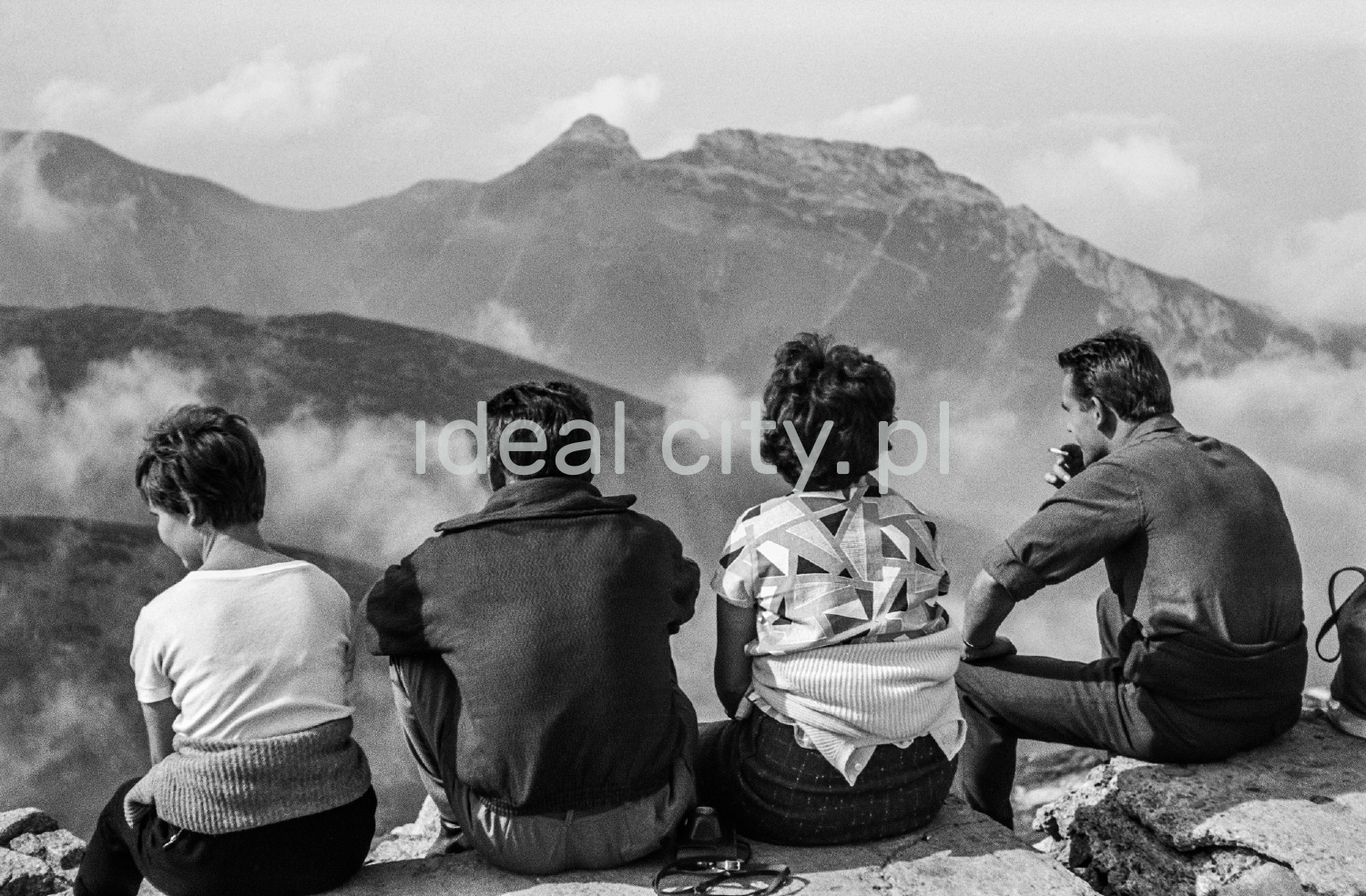 Tourists are resting on a rocky trail with a view of the Tatra peaks in the background.