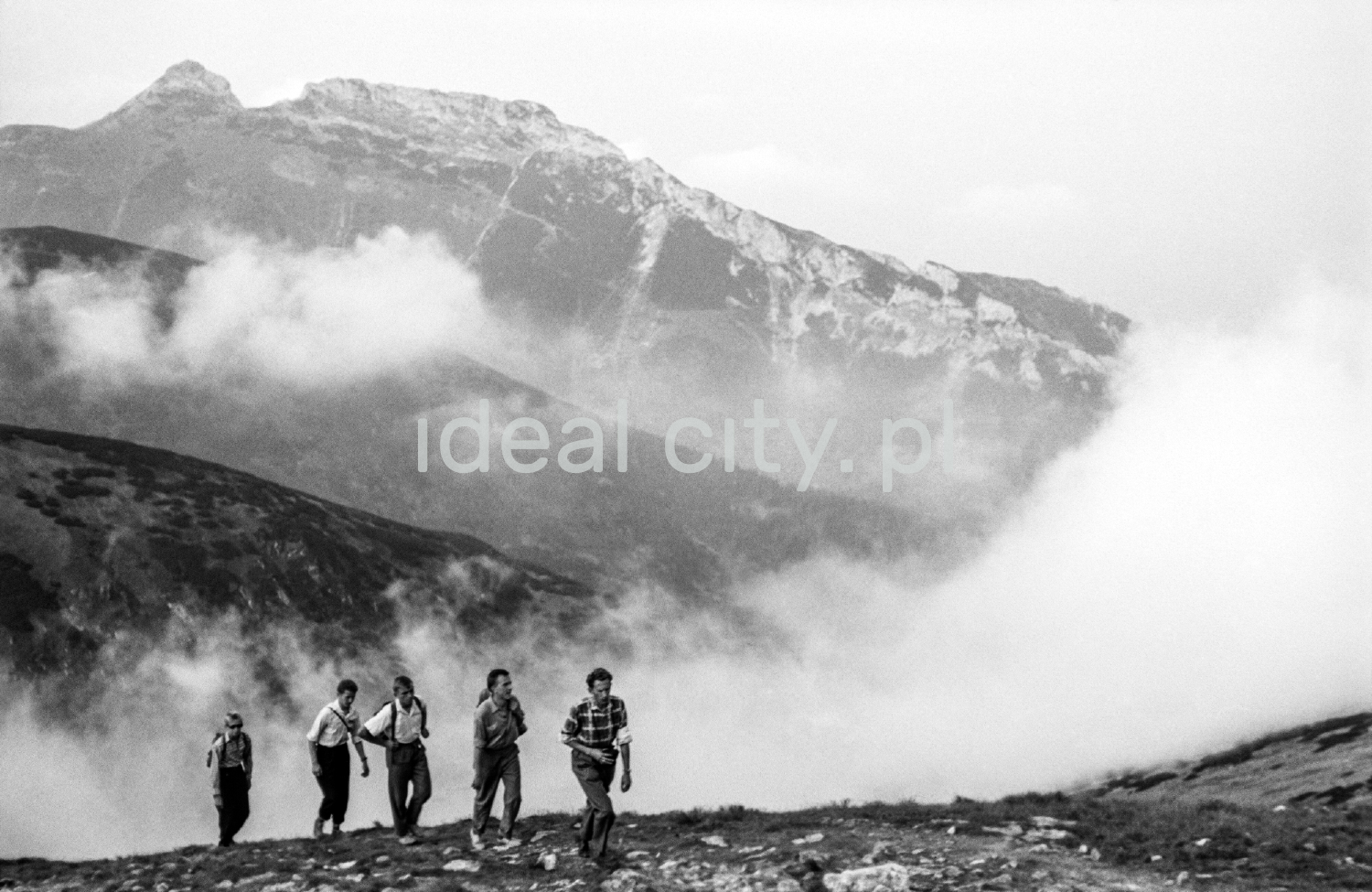Tourists with backpacks are walking along the rocky trail, with the perspective of the Tatra peaks in the background.