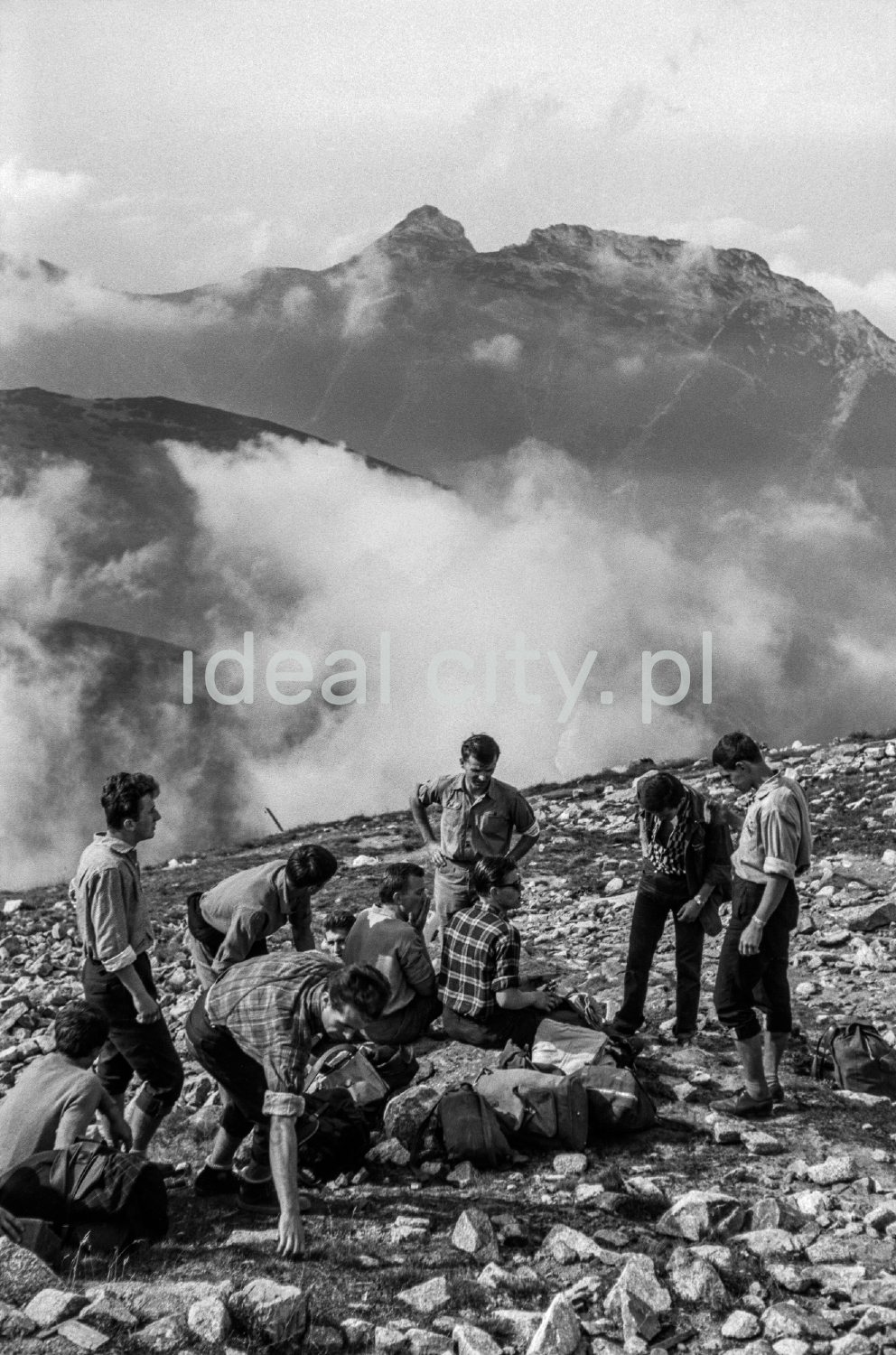 Tourists with backpacks are resting on a rocky trail with a view of the Tatra peaks in the background.