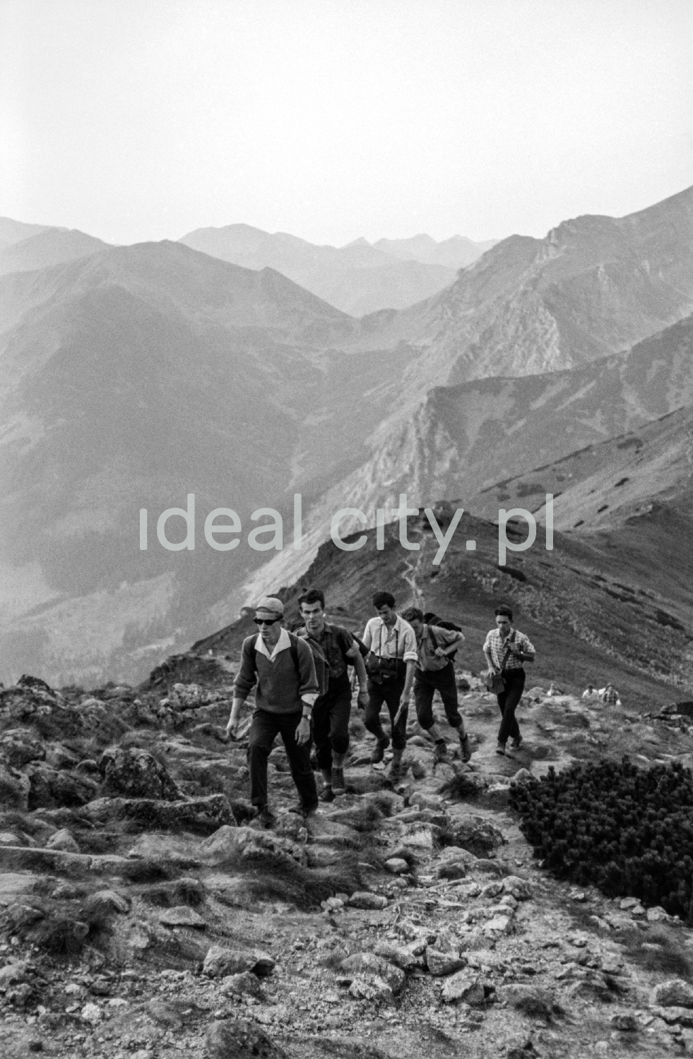 Tourists with backpacks are walking along the rocky trail, with the perspective of the Tatra peaks in the background.