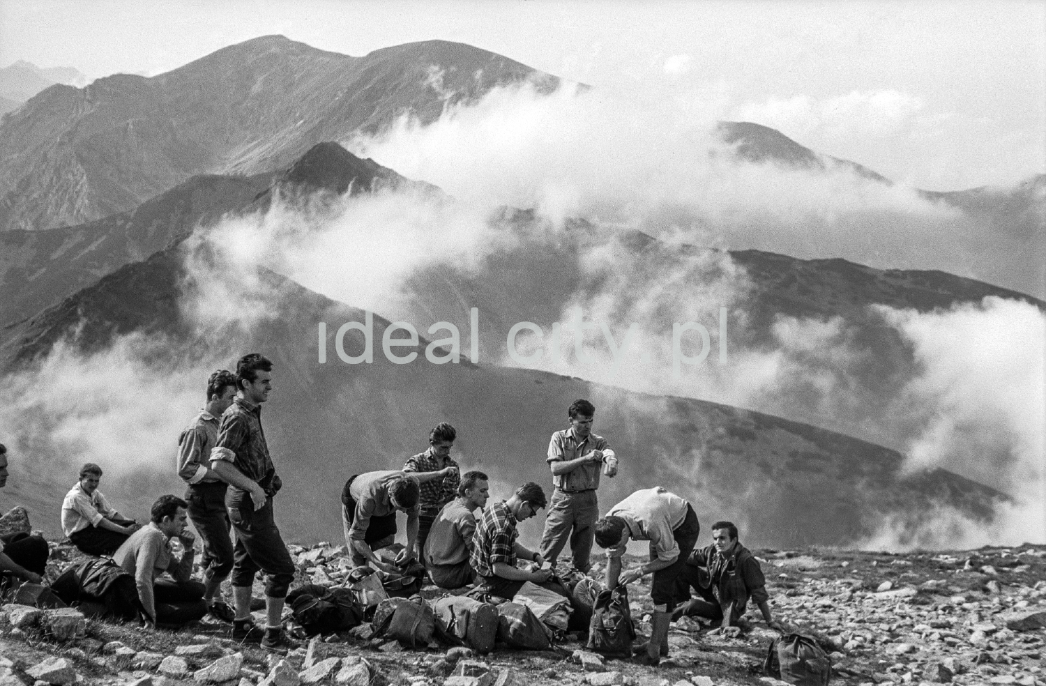 Tourists with backpacks are resting on a rocky trail with a view of the Tatra peaks in the background.