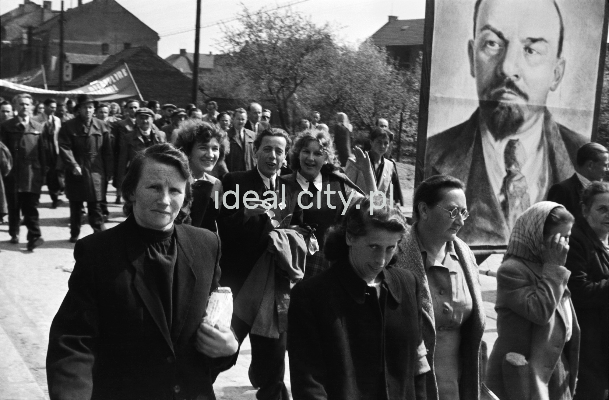 Odświętnie ubrani ludzie idą dynamicznie w pochodzie, osoby na pierwszym planie niosą portret Lenina.
