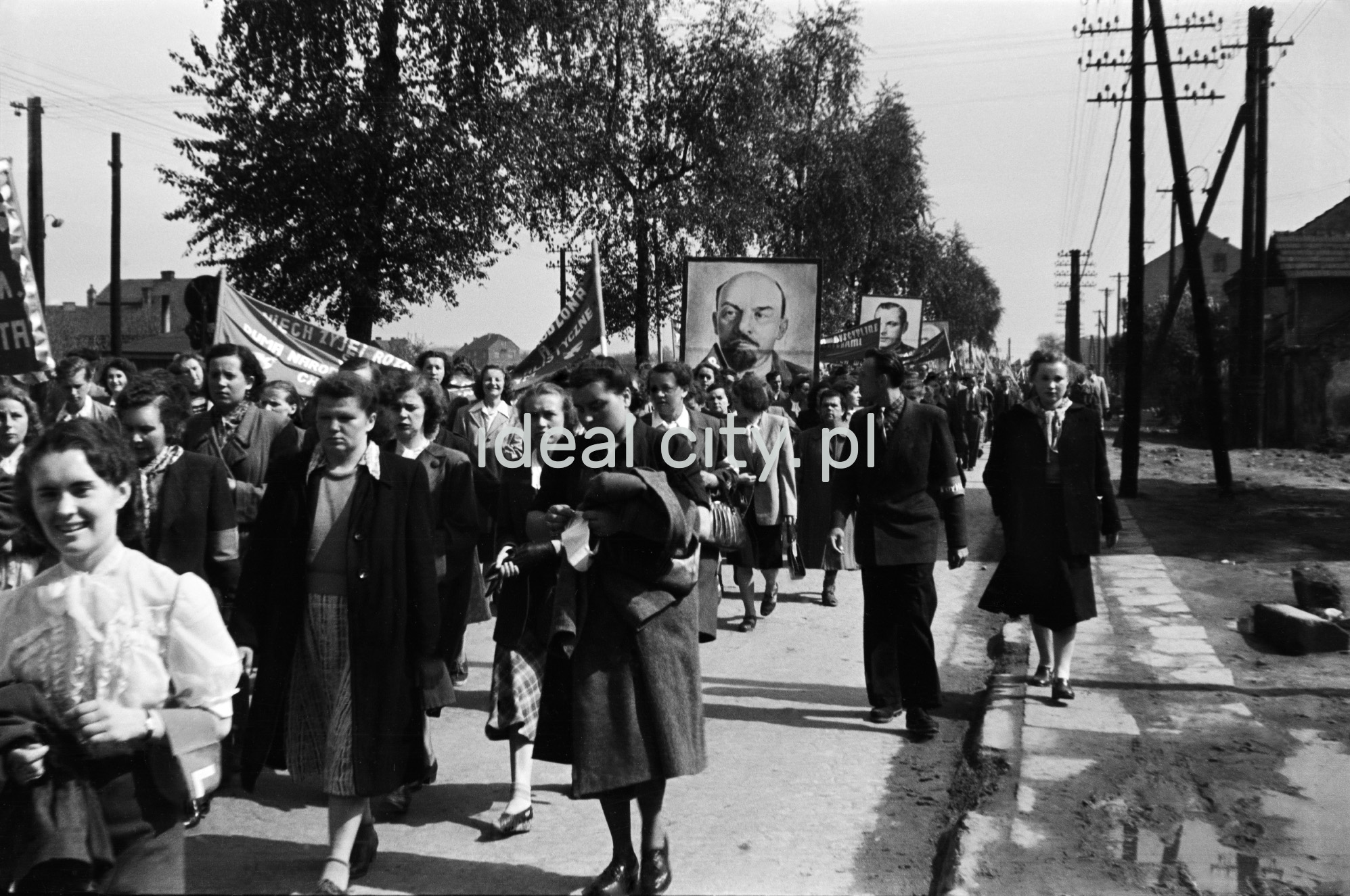 Odświętnie ubrani ludzie idą dynamicznie w pochodzie, w perspektywie ktoś niesie portret Lenina.