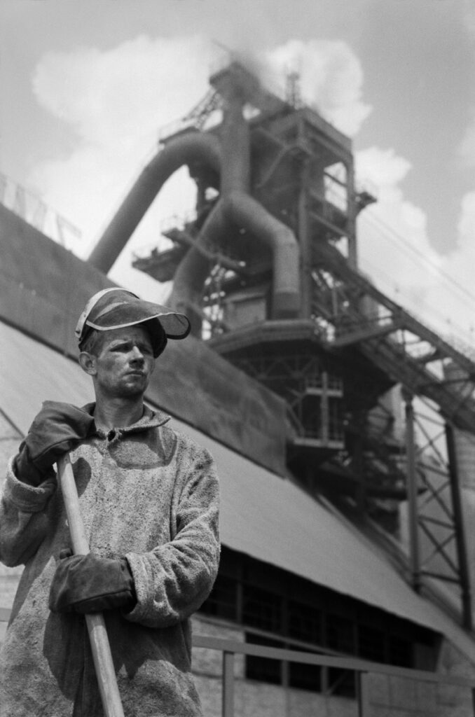 A portrait of a steelworker with work clothes in front of the steelworks building.