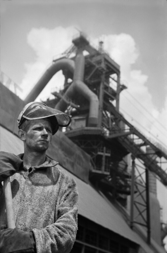 A portrait of a steelworker with work clothes in front of the steelworks building.