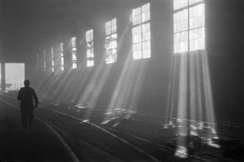 The factory hall is illuminated by streams of light, between them a figure in workwear