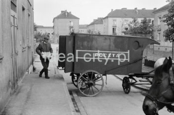 Pocztowy wóz konny do przewożenia paczek i listów przed budynkiem pierwszej poczty w Nowej Hucie na osiedlu A-1 Północ (Willowe). Została ona otwarta w połowie 1951 roku. Wystrój wnętrza, m.in. meble, kolumny pokryte marmurową okładziną, marmurowe blaty, zaprojektował Marian Sigmund. Pierwotnie w tym budynku (na piętrze) miał swoją siedzibę również Międzyzwiązkowy Klub Robotniczy. Na parterze znajdowały się punkty przyjęcia klientów, a na piętrze m.in. rozdzielnia oraz mieszkanie służbowe listonosza, który był jednocześnie stróżem. Listonosze dzielili się na miejskich i wiejskich, norma listonosza miejskiego wynosiła 2 tysiące mieszkańców. Na początku pracowało 7 listonoszy. W tle widoczne domy mieszkalne według projektów warszawskiego architekta Franciszka Adamskiego. Lata 50/60. XXw.

fot. Wiktor Pental/idealcity.pl

