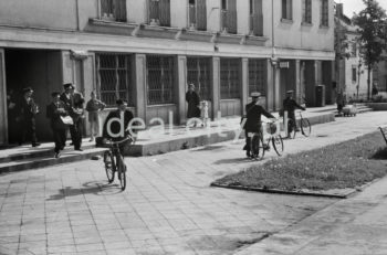 Listonosze wyprowadzający rowery z budynku poczty na osiedlu A-1 Północ (Willowe). Poczta została otwarta w połowie 1951 roku. Wystrój wnętrza, m.in. meble, kolumny pokryte marmurową okładziną, marmurowe blaty, zaprojektował Marian Sigmund. Pierwotnie w tym budynku (na piętrze) miał swoją siedzibę również Międzyzwiązkowy Klub Robotniczy. Na parterze znajdowały się punkty przyjęcia klientów, a na piętrze m.in. rozdzielnia oraz mieszkanie służbowe listonosza, który był jednocześnie stróżem. Listonosze dzielili się na miejskich i wiejskich, norma listonosza miejskiego wynosiła 2 tysiące mieszkańców. Na początku pracowało 7 listonoszy. W tle widoczne domy mieszkalne według projektów warszawskiego architekta Franciszka Adamskiego. Koniec lat 50. XXw.

fot. Wiktor Pental/idealcity.pl

