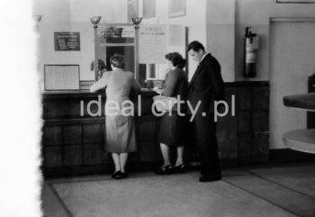 Wnętrze pierwszego urzędu pocztowego w Nowej Hucie, na osiedlu A-1 Północ (Willowe). Poczta została otwarta w połowie 1951 roku. Wystrój wnętrza, m.in. meble, kolumny pokryte marmurową okładziną, marmurowe blaty, zaprojektował Marian Sigmund. Pierwotnie w tym budynku (na piętrze) miał swoją siedzibę również Międzyzwiązkowy Klub Robotniczy. Na parterze znajdowały się punkty przyjęcia klientów, a na piętrze m.in. rozdzielnia oraz mieszkanie służbowe listonosza, który był jednocześnie stróżem. Listonosze dzielili się na miejskich i wiejskich, norma listonosza miejskiego wynosiła 2 tysiące mieszkańców. Na początku pracowało 7 listonoszy. W tle widoczne domy mieszkalne według projektów warszawskiego architekta Franciszka Adamskiego. Koniec lat 50. XXw. 

fot. Wiktor Pental/idealcity.pl

