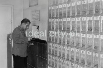 Wnętrze pierwszego urzędu pocztowego w Nowej Hucie, na osiedlu A-1 Północ (Willowe). Poczta została otwarta w połowie 1951 roku. Wystrój wnętrza, m.in. meble, kolumny pokryte marmurową okładziną, marmurowe blaty, zaprojektował Marian Sigmund. Pierwotnie w tym budynku (na piętrze) miał swoją siedzibę również Międzyzwiązkowy Klub Robotniczy. Na parterze znajdowały się punkty przyjęcia klientów, a na piętrze m.in. rozdzielnia oraz mieszkanie służbowe listonosza, który był jednocześnie stróżem. Listonosze dzielili się na miejskich i wiejskich, norma listonosza miejskiego wynosiła 2 tysiące mieszkańców. Na początku pracowało 7 listonoszy. W tle widoczne domy mieszkalne według projektów warszawskiego architekta Franciszka Adamskiego. Koniec lat 50. XXw. 

fot. Wiktor Pental/idealcity.pl

