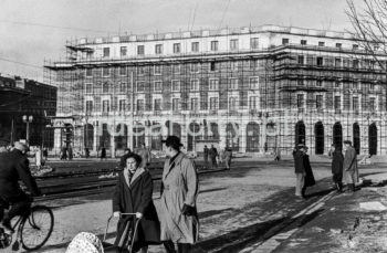 Północna pierzeja Placu Centralnego, widok w kierunku osiedla B-31 (Centrum B) i wylotu alei Lenina (obecnie Solidarności), budynek mieszkalny z lokalami usługowymi na parterze z rusztowaniami podczas tynkowania fasady.  Później od prawej strony, pod arkadami znajdował się na parterze sklep obuwniczy. Projektantem żyrandoli, mebli oraz gablot wystawienniczych był Marian Steczowicz wspólnie z Barbarą Gołajewską. Obok znajdował się sklep z materiałami tekstylnymi „Merino” z wielkimi lustrami, ozdobnymi żyrandolami i masywnymi meblami. Wnętrze zaprojektował Zdzisław Szpyrkowski. Kolejnym lokalem użytkowym był sklep jubilerski z kryształowymi żyrandolami. Projektantem wystroju wnętrza był Czesław Wallis (?). Za nim umiejscowiony był sklep Centrali Przemysłu Ludowego i Artystycznymi „Cepelia”. Na szczególna uwagę zasługiwały w nim: metalowe żyrandole z ręcznie malowanymi elementami ceramicznymi, kolorowe talerze ceramiczne umieszczone w kasetonach oraz uzupełniające całość wystroju wnętrza masywne meble. Elementy i ozdoby ceramiczne wykonała Spółdzielnia rzemiosła Artystycznego „Kamionka” z Łysej Góry, a meble Spółdzielnia Rękodzieła Artystycznego „Meble Artystyczne