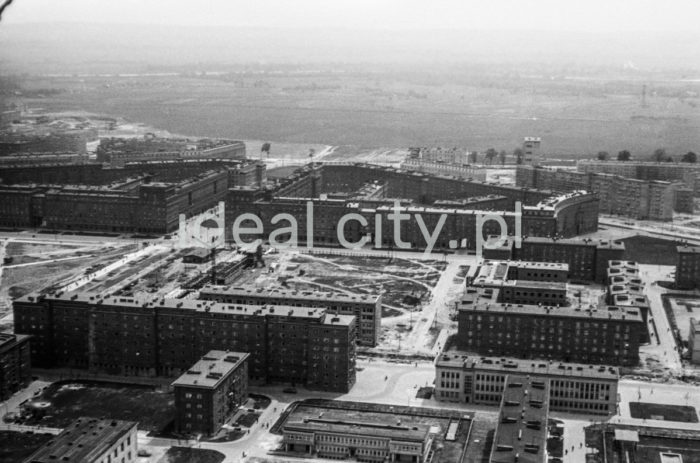 An aerial photograph depicting a view from Zone C estates: Urocze (C-33) and Zgody (C-32) Estates, towards the central estates in Nowa Huta. In the foreground: buildings on the Urocze Estate, built from 1955 to 1958. Ten out of the nineteen buildings were designed by architect Jan Suliga, who also designed small architecture for some of them. On the estate there is also an inoperative fountain. Behind: Zgody Estate with the former seat of the District Militia Station in Nowa Huta (now Police Station). In the empty square beginning construction works of a modernist building, the characteristic building known as “Świat Dziecka” (Child’s World – the name of a shop and its neon sign). It was built in 1957-60.
The semi-circular residential block no 7 on the corner is a characteristic feature of this estate. Behind it: Centrum D (D-31) Estate with the characteristic modernist tower block known as “helicopter.” Left: buildings on the Centrum B (B-31) and Centrum A (A-31) Estates, built in 1953-6, further back: green areas known as Nowa Huta Meadows. Late 1950s.

Fotografia lotnicza przedstawia widok od strony osiedli w strefie C: osiedla Uroczego (C-33) Zgody (C-32) w kierunku nowohuckich osiedli centralnych. Na pierwszym planie zabudowania na osiedlu Uroczym, realizowane było w latach 1955-1958. Dziesięć z dziewiętnastu budynków zaprojektował arch. Jan Suliga, który był także projektantem małej architektury przy kilku z nich. Wewnątrz osiedla znajduje się także nieczynna fontanna.  Kolejne, to osiedle Zgody z siedzibą dawnej Komendy Dzielnicowej MO w Nowej Hucie (obecnie mieści się w nim Komisariat Policji). Na pustym placu początki prac budowlanych przy modernistycznym gmachu charakterystyczny budynek zwany „Światem Dziecka” (od nazwy sklepu i neonu). Wzniesiono go w latach 1957-1960.

Na tym osiedlu charakterystyczny jest narożny blok mieszkalny nr 7 zbudowany na planie fragmentu koła. Za nim widoczne osiedl Centrum D (D-31) z charakterystycznym modernistycznym punktowcem zwanym „helikopterem”. Po lewej stronie widoczna zabudowa osiedli Centrum B (B-31)  i Centrum A (A-31) zrealizowanymi w latach 1953-1956, w tle widoczne tereny zielone zwane „Łąkami nowohuckimi”, koniec l.50.XX w.

Photo by Henryk Makarewicz/idealcity.pl

