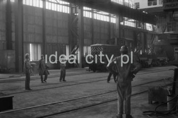 Lenin Metallurgical Combine, inside Converter Plant, drawing a ladle. Second half of the 1960s.
The Converter Plant was opened in February 1966. It was the first steel factory of that kind in Poland. The main building housed the converter hall with two converters, a charging bay and two casting halls. The charge consisted of 70% molten pig iron and about 30% scrap. Molten pig iron came from mixers on torpedo cars holding ladles. Steel was cast into casting ladles placed on a power-driven steel car that crossed casting halls. Once ingot moulds were taken off in their destination hall, ingots were transferred to soaking pits in preparatory rolling mills. Slag was carried to slag disposal sites by slag cars.

Kombinat metalurgiczny im. Lenina, wnętrze hali Stalowni Konwertorowej, wyciąganie kadzi, II połowa lat 60.
Stalownię uruchomiono w lutym 1966 roku. Była pierwszą tego typu stalownią w Polsce. Budynek główny obejmował halę konwerterów z dwoma konwertorami, halę wsadu oraz dwie hale odlewnicze. Wsad metaliczny stanowił 70% płynnej surówki i ok. 30% złomu. Surówka ciekła dostarczana była z mieszalników na surówkowozach, wyposażonych w kadzie. Odlewanie stali odbywało się do kadzi odlewniczych, umieszczonych na napędzanym elektrycznie wozie stalowniczym, jeżdżącym w poprzek hal odlewniczych. Po ściągnięciu wlewnic w odpowiedniej hali, wlewki w stanie gorącym przekazywano do pieców wgłębnych walcowni wstępnych. Żużel wywożono do kafaru na żużlowozach.

Photo by Henryk Makarewicz/idealcity.pl

