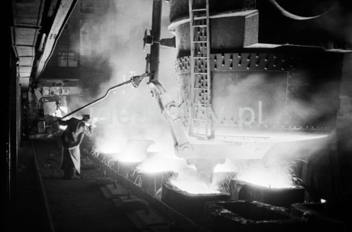 Lenin Metallurgical Combine, inside Converter Plant, pouring steel into ingot moulds. Second half of the 1960s.
The Converter Plant was opened in February 1966. It was the first steel factory of that kind in Poland. The main building housed the converter hall with two converters, a charging bay and two casting halls. The charge consisted of 70% molten pig iron and about 30% scrap. Molten pig iron came from mixers on torpedo cars holding ladles. Steel was cast into casting ladles placed on a power-driven steel car that crossed casting halls. Once ingot moulds were taken off in their destination hall, ingots were transferred to soaking pits in preparatory rolling mills. Slag was carried to slag disposal sites by slag cars.

Kombinat metalurgiczny im. Lenina, wnętrze hali Stalowni Konwertorowej, rozlewanie stali do wlewnic.
Stalownię uruchomiono w lutym 1966 roku. Była pierwszą tego typu stalownią w Polsce. Budynek główny obejmował halę konwerterów z dwoma konwertorami, halę wsadu oraz dwie hale odlewnicze. Wsad metaliczny stanowił 70% płynnej surówki i ok. 30% złomu. Surówka ciekła dostarczana była z mieszalników na surówkowozach, wyposażonych w kadzie. Odlewanie stali odbywało się do kadzi odlewniczych, umieszczonych na napędzanym elektrycznie wozie stalowniczym, jeżdżącym w poprzek hal odlewniczych. Po ściągnięciu wlewnic w odpowiedniej hali, wlewki w stanie gorącym przekazywano do pieców wgłębnych walcowni wstępnych. Żużel wywożono do kafaru na żużlowozach.

Photo by Henryk Makarewicz/idealcity.pl

