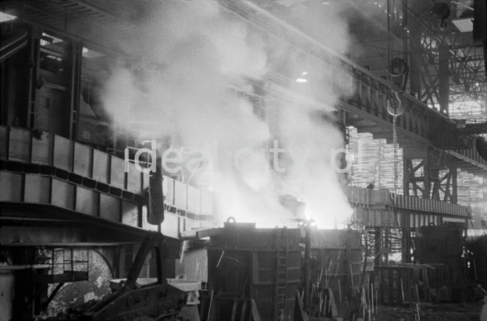 Lenin Metallurgical Combine, inside Converter Plant, pouring steel into ingot moulds. Second half of the 1960s.
The Converter Plant was opened in February 1966. It was the first steel factory of that kind in Poland. The main building housed the converter hall with two converters, a charging bay and two casting halls. The charge consisted of 70% molten pig iron and about 30% scrap. Molten pig iron came from mixers on torpedo cars holding ladles. Steel was cast into casting ladles placed on a power-driven steel car that crossed casting halls. Once ingot moulds were taken off in their destination hall, ingots were transferred to soaking pits in preparatory rolling mills. Slag was carried to slag disposal sites by slag cars.

Kombinat metalurgiczny im. Lenina, wnętrze hali Stalowni Konwertorowej, rozlewanie stali do wlewnic.
Stalownię uruchomiono w lutym 1966 roku. Była pierwszą tego typu stalownią w Polsce. Budynek główny obejmował halę konwerterów z dwoma konwertorami, halę wsadu oraz dwie hale odlewnicze. Wsad metaliczny stanowił 70% płynnej surówki i ok. 30% złomu. Surówka ciekła dostarczana była z mieszalników na surówkowozach, wyposażonych w kadzie. Odlewanie stali odbywało się do kadzi odlewniczych, umieszczonych na napędzanym elektrycznie wozie stalowniczym, jeżdżącym w poprzek hal odlewniczych. Po ściągnięciu wlewnic w odpowiedniej hali, wlewki w stanie gorącym przekazywano do pieców wgłębnych walcowni wstępnych. Żużel wywożono do kafaru na żużlowozach.

Photo by Henryk Makarewicz/idealcity.pl


