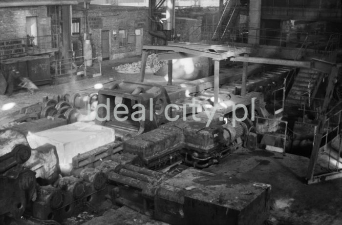 Lenin Metallurgical Combine, inside the Zgniatacz Rolling Mill, rolling ingots no. 1470. 1960s.
The construction of the Zgniatacz Rolling Mill commenced as soon as 1953, first output was produced in 1955. Here carbon and low alloy steel ingots weighing between 8 and 15 tons were preliminarily processed into slabs or square blooms. Cast slabs are rolled at various thicknesses (100 to 200 mm), widths (550-1550 mm) and lengths (1250-1550 mm). Cold or hot ingots were transported from the steel plant by railway to the soaking pits hall, where they were heated to a temperature of about 1300˚C. The blooming mill (zgniatacz) was a duo reversible rolling mill. Each roller was driven by an individual electric engine. Hot blooms were sent directly to the billet rolling mill. Billets could then be moved directly to the finishing room or onto loading screens at the continuous steel strip rolling mill. 1960s.

Kombinat metalurgiczny im. Lenina, wnętrze hali Walcowni Zgniatacz, walcowanie wlewek nr 1470, lata 60.
Wstępne prace przy budowie Walcowni Zgniatacz rozpoczęto już w 1953 roku, produkcja nastąpiła w 1955 roku. To walcownia wstępnego przerobu wlewków ze stali węglowej i niskostopowej o ciężarze od 8 do 15 ton na kęsiska płaskie lub kwadratowe. Odlewane kęsiska płaskie walcowane są w różnych grubościach (od 100 do 200 mm), szerokościach (od 550 do 1550 mm) oraz długościach (od 1250 do 1550 mm). Wlewki w stanie zimnym lub gorącym dostarczano ze stalowni trakcją kolejową do hali pieców wgłębnych. Piece te służyły do nagrzewania wlewków do temperatury ok. 1300 ℃. Zgniatacz był walcarką duo-nawrotną. Każdy walec posiadał napęd indywidualny za pomocą silników elektrycznych. Kęsiska ze zgniatacza kierowane były w stanie gorącym bezpośrednio do walcowni kęsów. Pozwalała ona kierować kęsy płaskie bezpośrednio do wykańczalni lub na kraty załadowcze walcowni ciągłej taśm, lata 60.

Photo by Henryk Makarewicz/idealcity.pl


