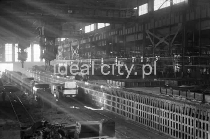 Kombinat metalurgiczny im. Lenina, wnętrze hali Walcowni Zgniatacz. Wstępne prace przy budowie Walcowni Zgniatacz rozpoczęto już w 1953 roku, produkcja nastąpiła w 1955 roku. To walcownia wstępnego przerobu wlewków ze stali węglowej i niskostopowej o ciężarze od 8 do 15 ton na kęsiska płaskie lub kwadratowe. Odlewane kęsiska płaskie walcowane są w różnych grubościach (od 100 do 200 mm), szerokościach (od 550 do 1550 mm) oraz długościach (od 1250 do 1550 mm). Wlewki w stanie zimnym lub gorącym dostarczano ze stalowni trakcją kolejową do hali pieców wgłębnych. Piece te służyły do nagrzewania wlewków do temperatury ok. 1300 ℃. Zgniatacz był walcarką duo-nawrotną. Każdy walec posiadał napęd indywidualny za pomocą silników elektrycznych. Kęsiska ze zgniatacza kierowane były w stanie gorącym bezpośrednio do walcowni kęsów. Pozwalała ona kierować kęsy płaskie bezpośrednio do wykańczalni lub na kraty załadowcze walcowni ciągłej taśm, lata 60. 

fot. Henryk Makarewicz/idealcity.pl



